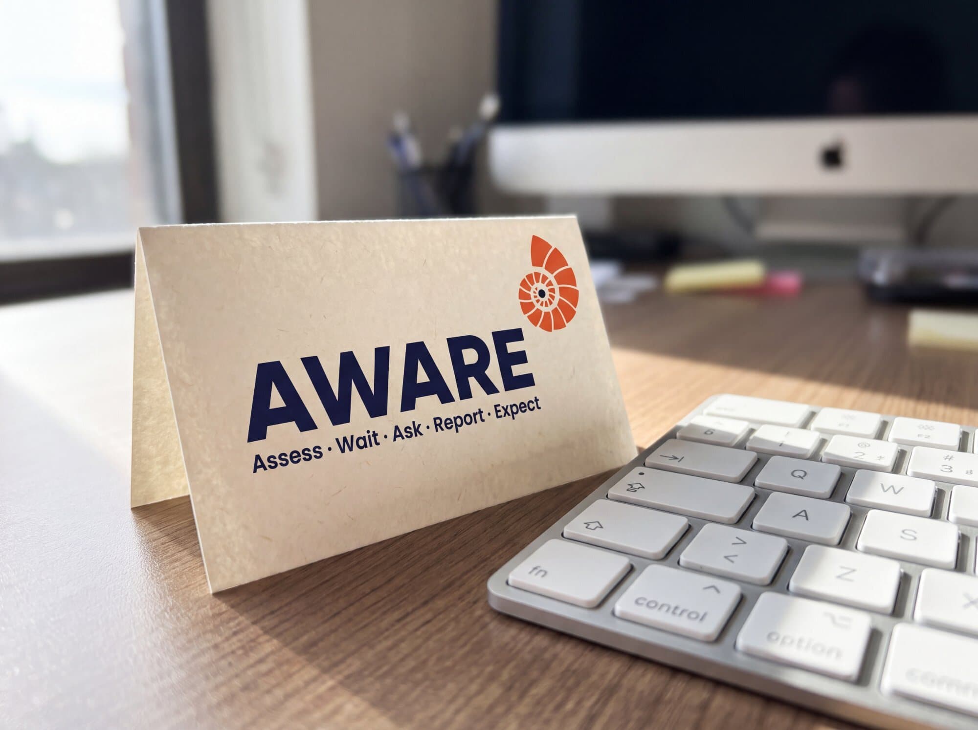 The AWARE desk card beside a keyboard: Assess, Wait, Ask, Report, Expect.